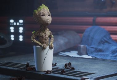 Baby Groot is planted in a cracked white pot and looking upward. The pot is on a workbench next to a wrench, and a blue tarp is in the background.