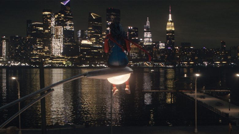 Iman Vellani as Ms. Marvel, aka Kamala Khan, sits atop a glowing streetlight as she surveys Manhattan in the distance
