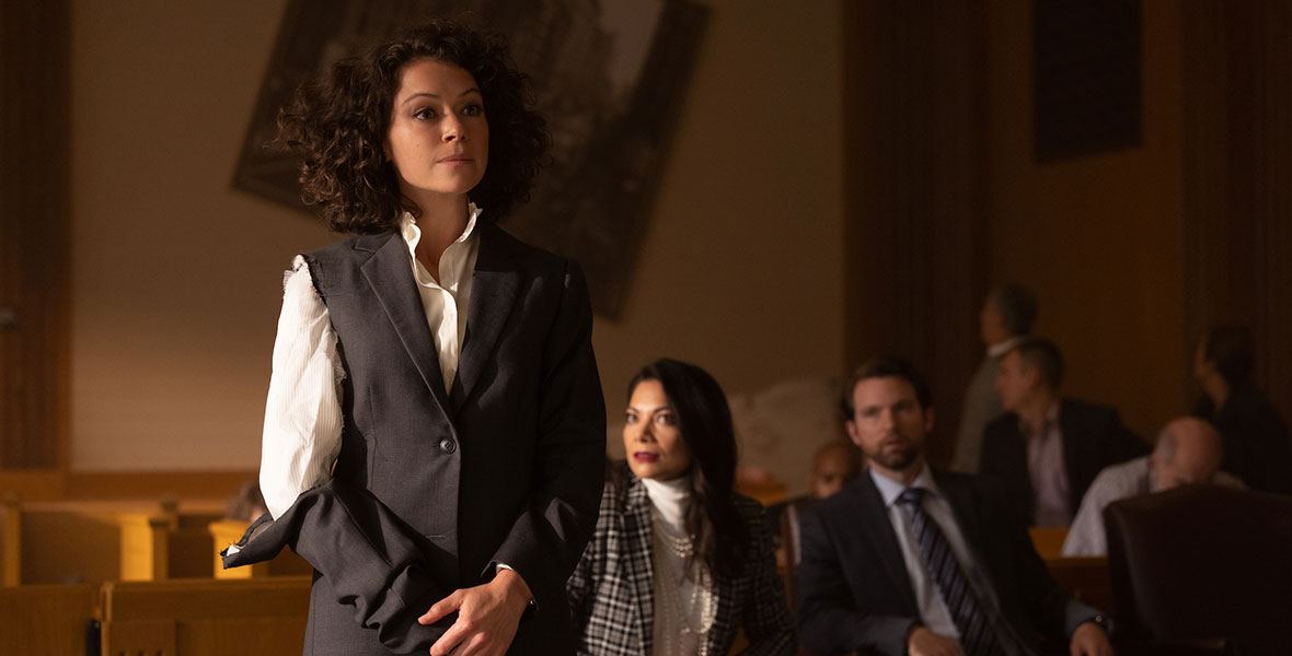 Actor Tatiana Maslany stands in a courtroom with one sleeve of her dark grey suit jacket ripped, exposing her white blouse. Behind her Ginger Gonzaga and Josh Segarra are seated in wooden chairs and a large portrait hangs askew.