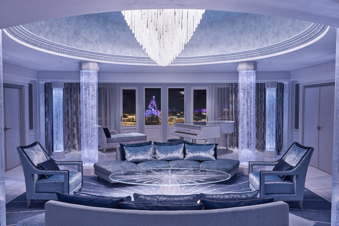 The Frozen Signature Suite has a chandelier hanging above a round table. The room has a silvery blue color scheme. Sleeping Beauty Castle is seen through the window.