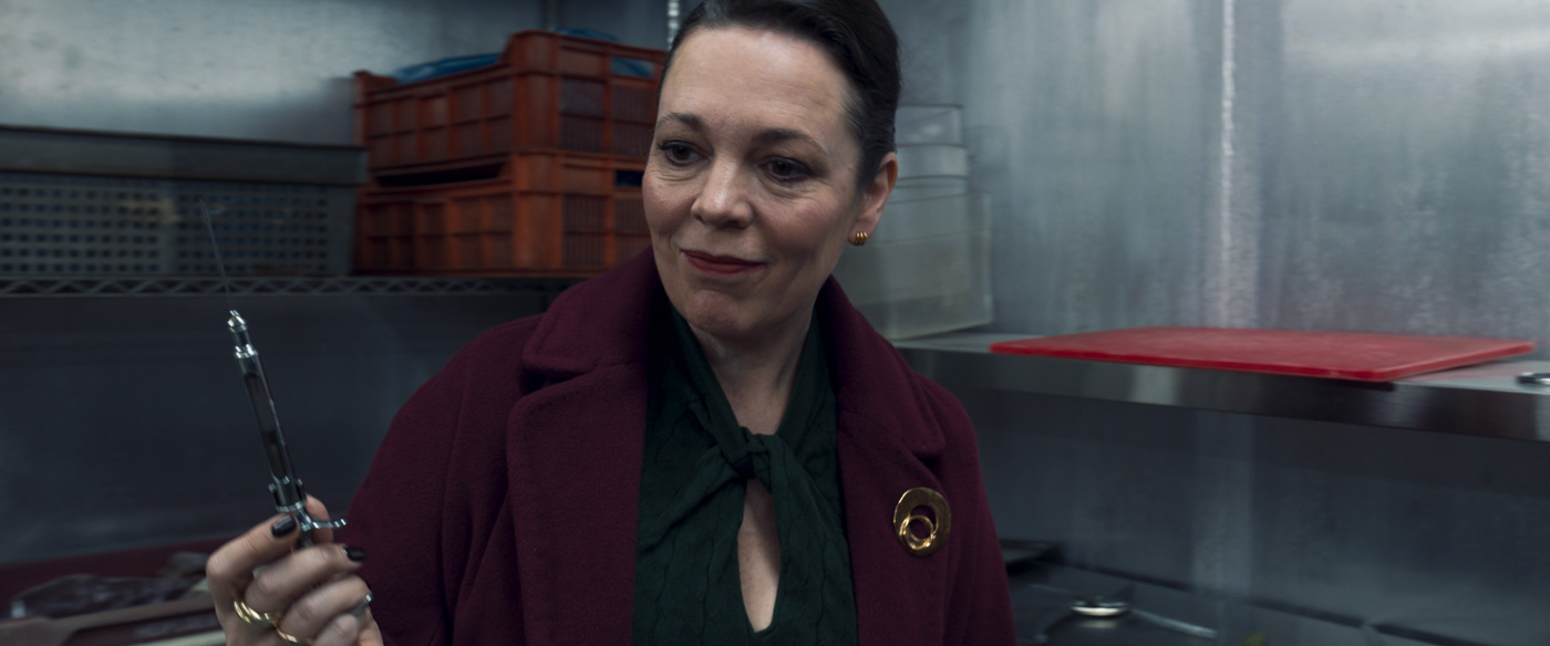 In a scene from the Marvel Studios series Secret Invasion, Sonya Falsworth, played by Olivia Colman, is holding a syringe with a needle attached to it. She is wearing a burgundy coat and black shirt.  