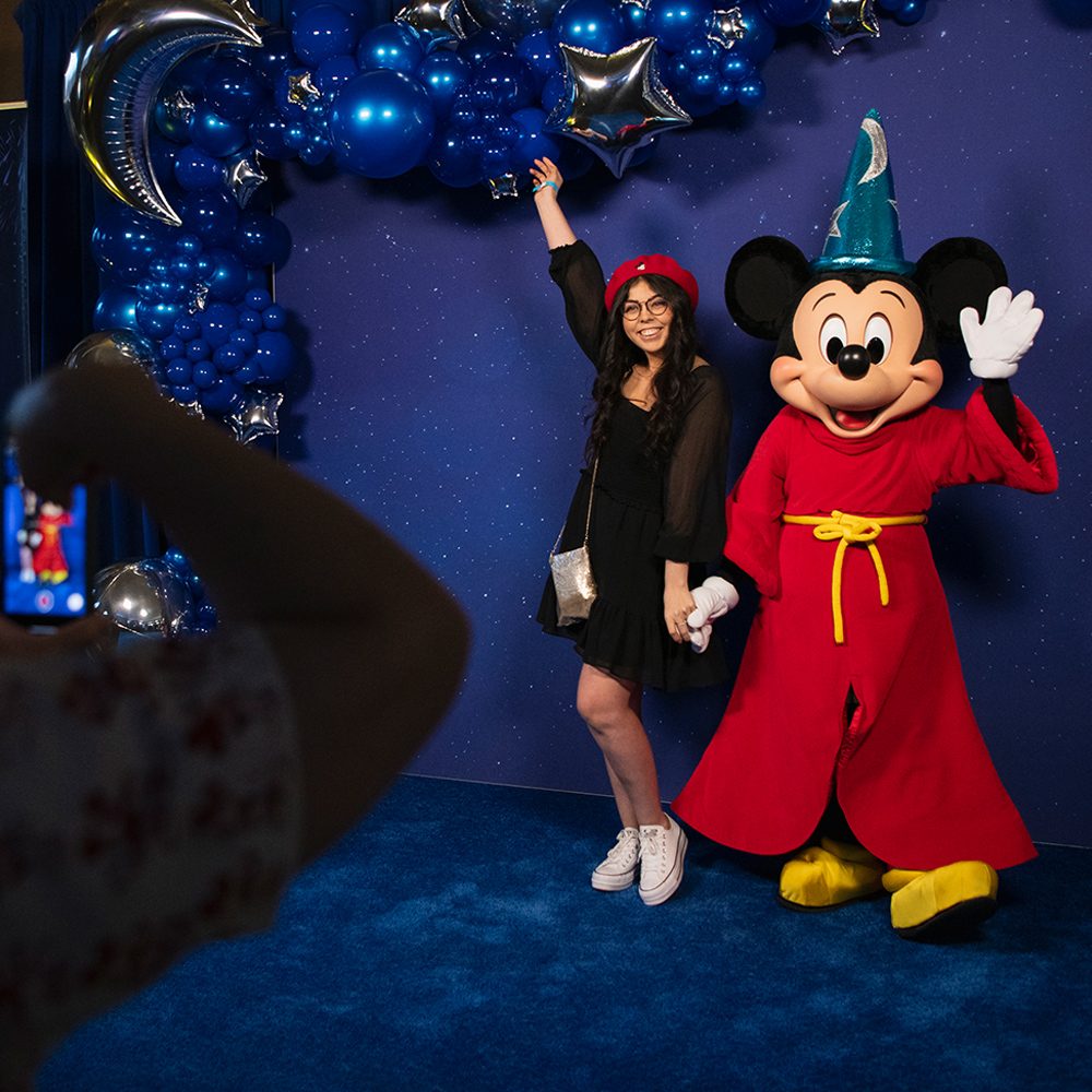 D23 Event A female D23 Member poses with Sorcerer's Apprentice Mickey Mouse in front of a starry sky back drop.