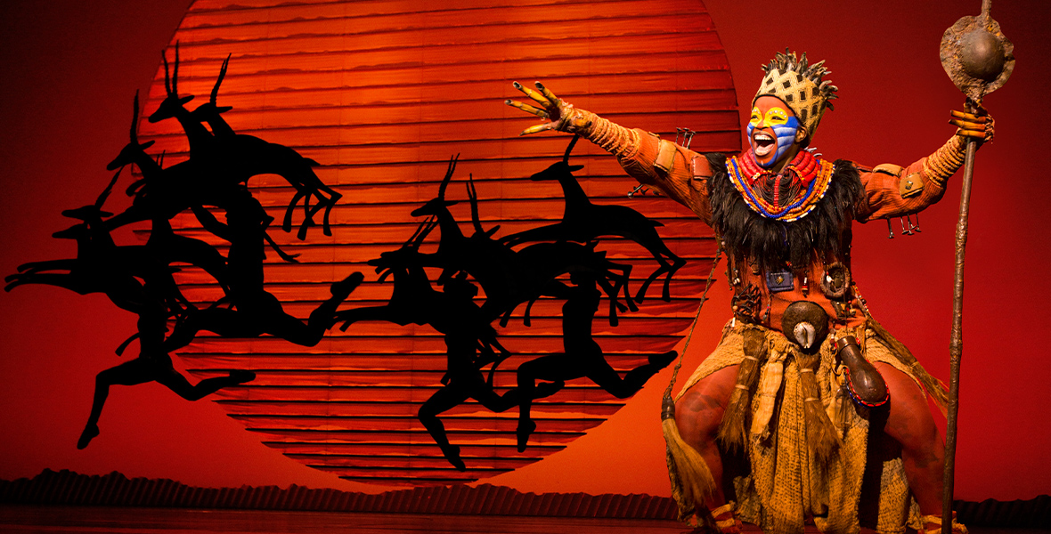 The Lion King Actor Mukelisiwe Goba performs on stage as Rafiki in Disney Theatricals’ The Lion King, with a large orange sun and silhouette of antelopes in the background. Goba is singing with power and holding their hand to the sky.