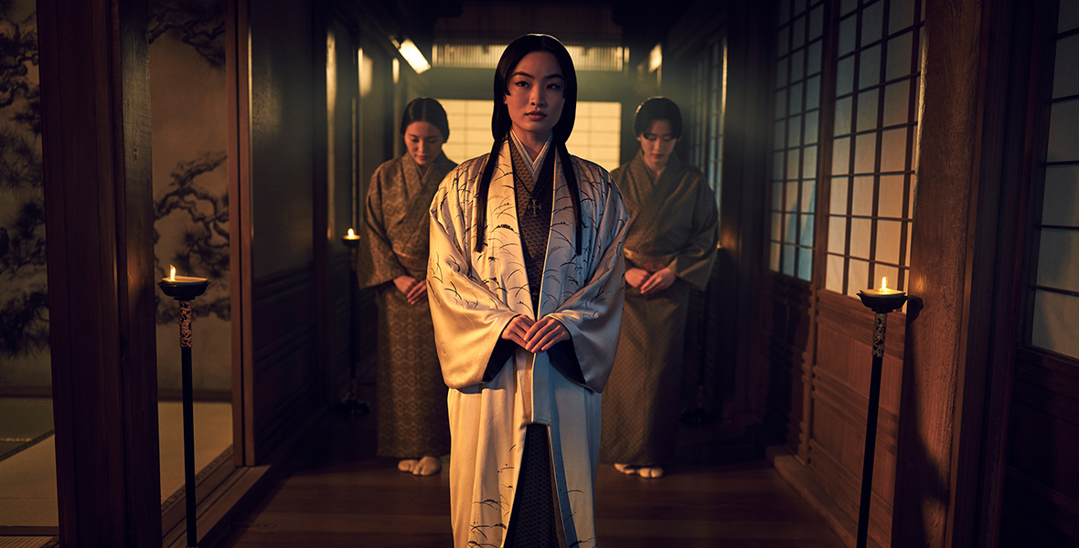 wears a white silk robe with black details over a brown kimono with orange patterns. Her long black hair falls forward in two strands, and she looks to her right. Two women beside her wear brown kimonos with patterns, their heads slightly bent forward in respect. This scene unfolds within a candlelit, Japanese-style hallway characterized by wooden flooring and traditional windows.