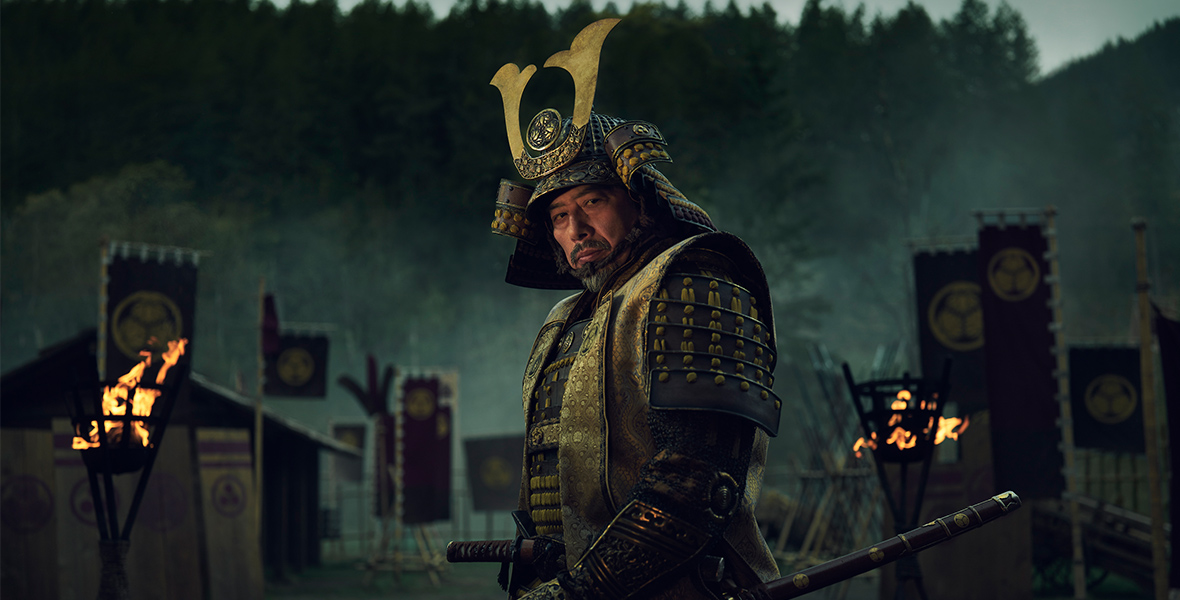 In a promotional image for FX’s Shōgun, Hiroyuki Sanada, portrayed by Yoshii Toranga, stands at the center of the image wearing traditional Japanese armor. Behind him, a lush forest and army tents create the backdrop, while sporadic torches cast an eerie, mysterious glow.