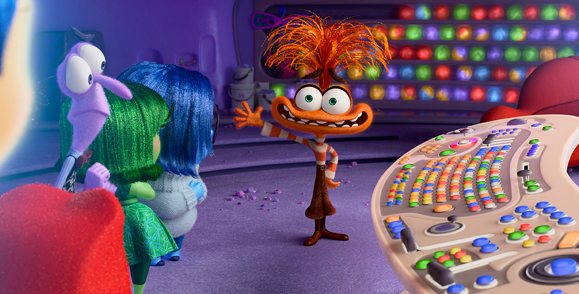 In a scene from Inside Out 2, Anxiety (voiced by Maya Hawke) stands facing the camera, nervously waving at the original Emotions lined up in front of her. To the right of the image is Headquarters' cream console with colorful buttons. Behind Anxiety, shelves display colorful marble balls. In front of them sits a red couch with orange pillows.