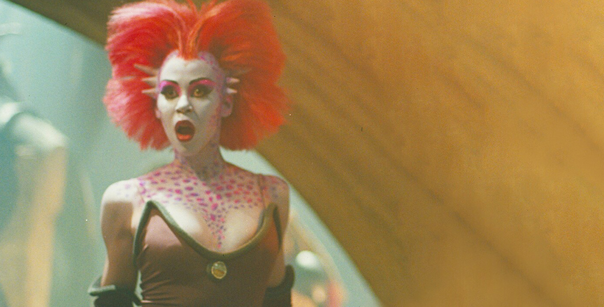 In an image from Star Wars Episode VI: Return of the Jedi, Rystáll Sant, a humanoid alien with bright red hair, spotted pink skin, and horns, gasps in shock at something happening offscreen.