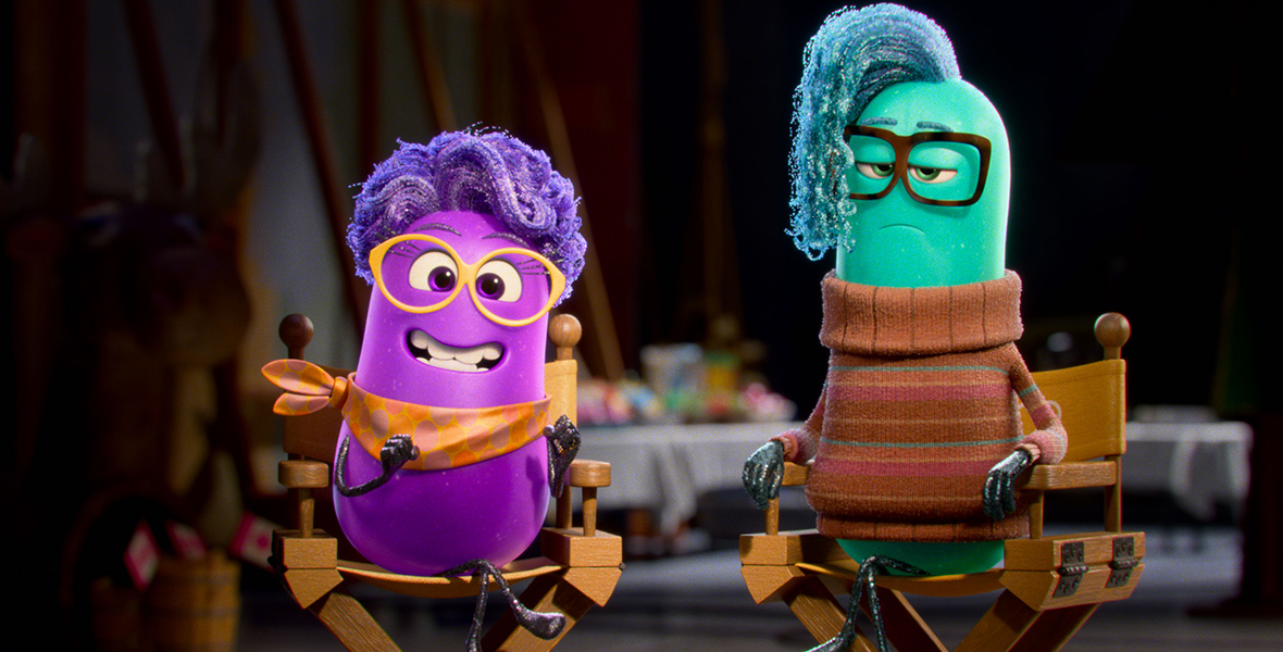 In an image from Dream Productions, Paula (voiced by Paula Pell), a purple, anthropomorphic, jellybean-like director, wears a purple wig, orange glasses, and an orange necktie as she smiles at the camera from her director’s chair. To her left is Xeni (voiced by Richard Ayoade), a turquoise, anthropomorphic, jellybean-like director with blue hair, brown glasses, and an orange striped turtleneck sweater. He also sits on a director’s chair. The background features a buffet of food inside a bustling soundstage.