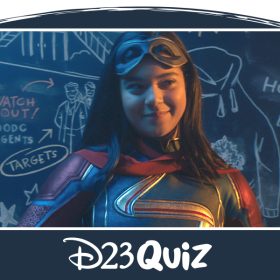 Kamala Khan, dressed as her hero alter ego Ms. Marvel, smiles while holding a piece of chalk. She is standing in front of a chalkboard with a variety of illustrations and plans drawn on it.