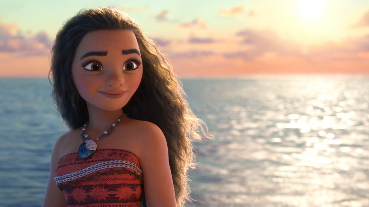 In a scene from Moana, Moana (voiced by Auliʻi Cravalho) smiles as she looks ahead. She wears a patterned strapless red top and a necklace. Her long, curly hair flows back in the wind, with the ocean and a vibrant sunset in the background.