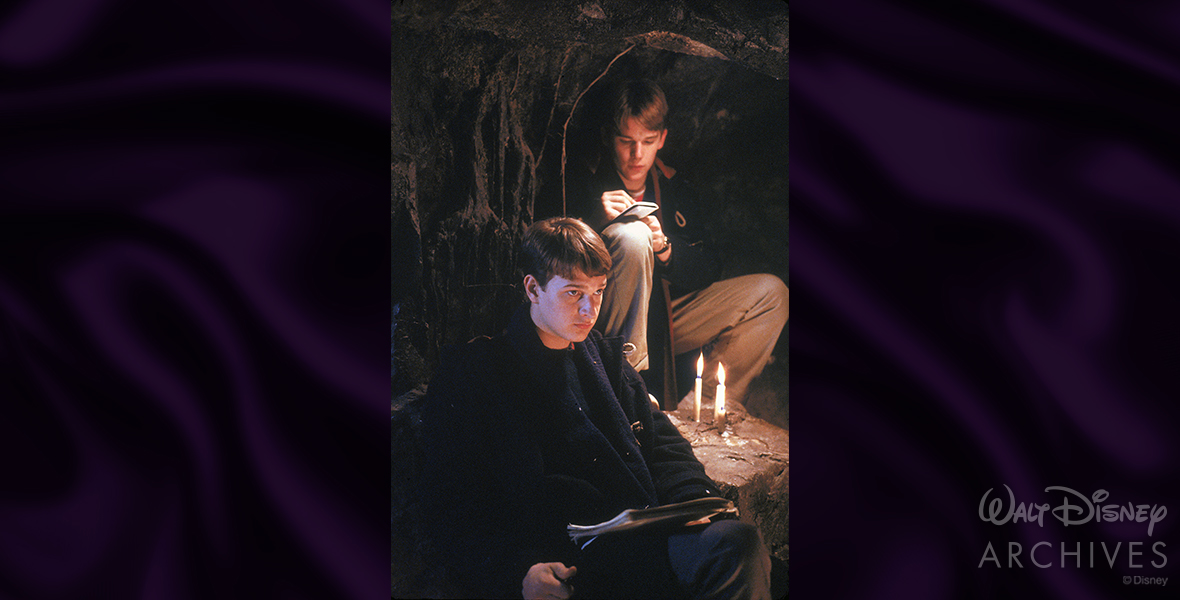 Josh Charles’ Knox Overstreet and Ethan Hawke’s Todd Anderson pose for a Dead Poets Society production photo while filming a scene inside the iconic cave. Charles on the bottom left wears a dark navy trench coat and looks forward, while Hawke wears khaki slacks and a dark navy trench coat and is seen writing in a small notebook. On a rock between the two are two lit candles.