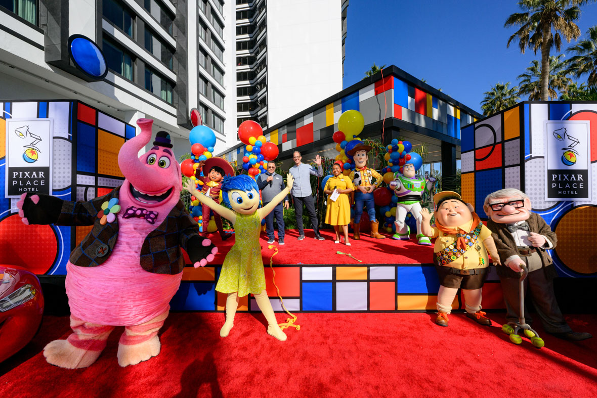 On a stage set up in front of Pixar Place Hotel at Disneyland Resort, a host of Pixar characters celebrate the hotel’s grand opening. The characters include Bing Bong and Joy from Inside Out, and Russell and Carl from Up in front of the stage. On the stage, along with Pixar Chief Creative Officer Pete Docter, are Miguel from Coco and Woody and Buzz from the Toy Story movies.