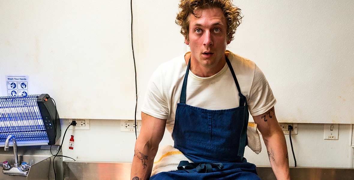 In a scene from the television show The Bear, Carmy, played by Jeremy White, is sitting on a metallic kitchen shelf. He is wearing a white T-shirt and a blue apron. Courtesy of FX Networks.