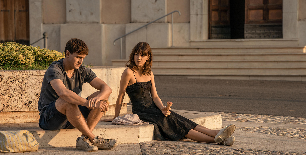 In a scene from the television series Normal People, Connell (left), played by Paul Mescal and Marianne (right), played by Daisy Edgar-Jones, are seated on a marble pavement. Connell wears a gray T-shirt, black cargo shorts, and sneakers, while Marianne wears a black midi dress and sneakers, holding an ice cream cone. Marianne gazes at Connell while he looks down.