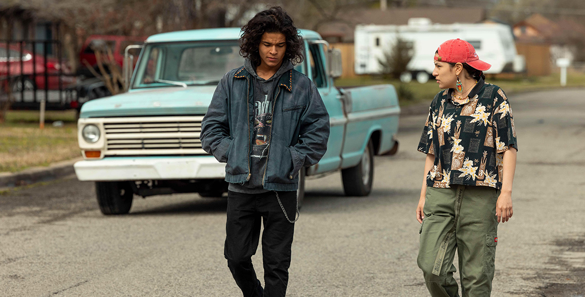In a scene from the television series Reservation Dogs, Bear Smallhill (left), played by D’Pharaoh Woon-A-Tai, is walking with Cheese (right), played by Lane Factor. Bear wears a gray graphic hoodie, a blue jacket, black jeans, and black sneakers. Cheese sports a red cap, a short-sleeve black shirt with floral details, green cargo pants, and black slides. In the background, a blue truck and a white trailer are visible.