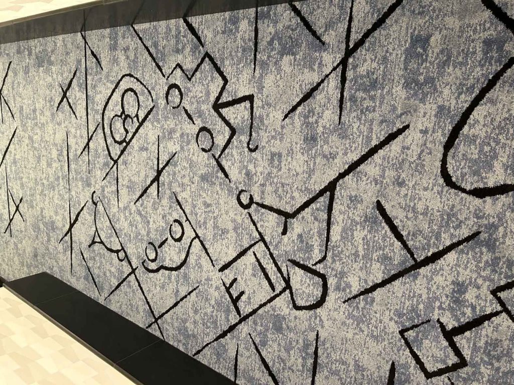 A gray rug in a hallway of Pixar Place Hotel. The rug features abstract line art in black of various Pixar characters, including Remy, Mater, and Edna Mode.