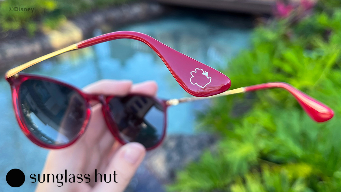 A pair of red sunglasses are held in hand in this close-up image. The focal point is on the red arms of the sunglasses, which are branded with the Minnie Mouse silhouette. 