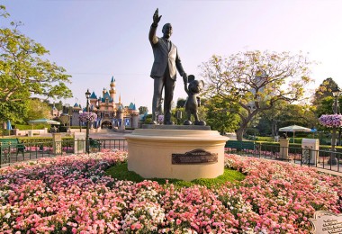 photo of statue of Walt Disney holding hands with Mickey Mouse at Disneyland, placard reading Partners, surrounded by large circular flower bed with Seeping Beauty Castle in background