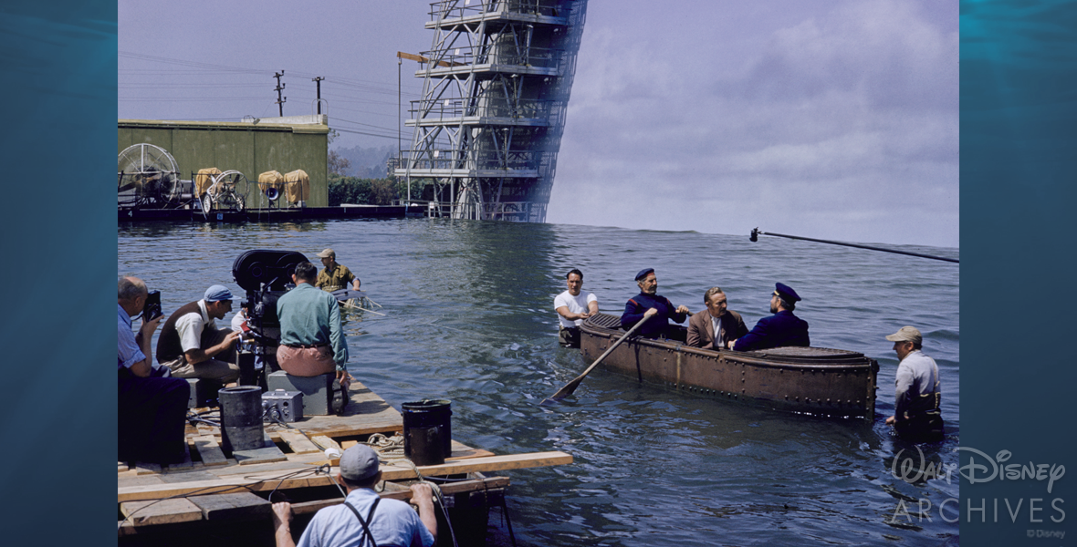 A rowboat is on the right of the photo floating on the dark blue waters of Sersen Lake. Professor Pierre Aronnax and Captain Nemo are face to face, having a conversation while another crew member of the Nautilus sits behind the professor, rowing. Two production crew members are on either side of the photo, positioning the small boat for filming. A boom mic, presumably held by something out of the frame, stretches above the actors. On the left of the photo, production crew members set up the camera for the scene. Behind the actors, the edge of the water seamlessly lines up with the artificially constructed cloudy backdrop. On the back left of the photo, wind machines can be seen on dry land as well as a green building.