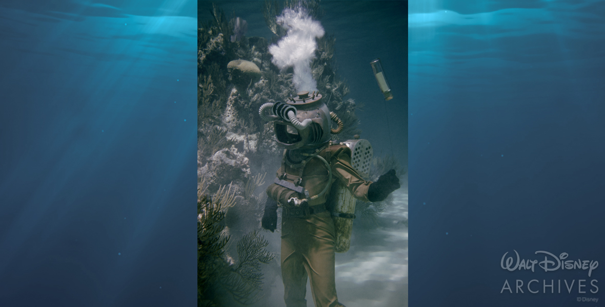 A crew member of the Nautilus stands on the bottom of the ocean. In his left hand, he grips a string tied to a message in a corked glass bottle. On the opposite side is a high gray mass of rock and coral. The crew member is wearing an intricate diving suit. It is a tan rubber jumpsuit with black gloves and a headpiece that resembles a Japanese pearl diving helmet. Strapped to his back is a breathing apparatus encased in silver metal. Above his head, air bubbles rush through the deep blue of the ocean toward the surface.