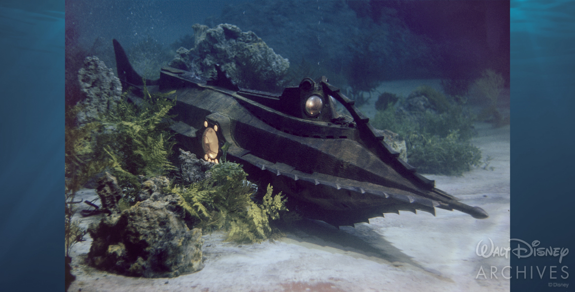A small scale model of the Nautilus is moving along what appears to be the bottom of the ocean. The full length of the gray spiked ship can be seen. A side porthole lit from within emits a warm yellow light. Shrubbery and rock formations are placed along both sides of the Nautilus.