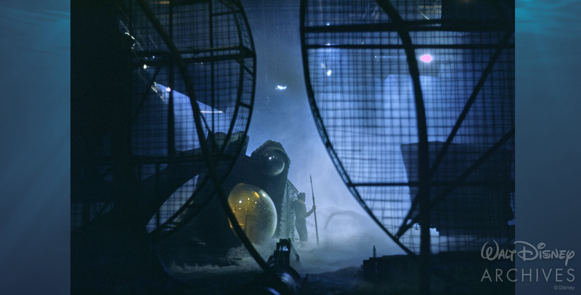 Two enormous wind machines are in the foreground, reaching from the bottom to the top of the photo. The cage-like exterior allows the viewer to look through them, seeing the battle between Captain Nemo and the giant squid. James Mason as Captain Nemo holds onto the side of the Nautilus; behind him is the yellow glowing eye of the ship. In his other hand, he holds onto a spear, ready to be launched at the squid. Only the tentacles of the squid can be seen; the rest of the body fades eerily into the fog of the water being strewn about by the wind machines to create this stormy battle.
